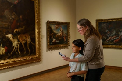 A parent holds their phone infront of a child, showing a guide to the European artwork they’re both standing infront of.