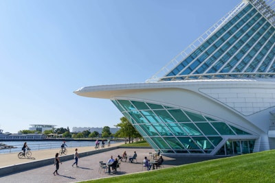 The Oak Leaf Trail to the right of Lake Michigan and the left of the Milwaukee Art Museum on a clear, sunny day. The trail and the museum’s Cafe patio are populated with cyclists, walkers, and guests drinking coffee.